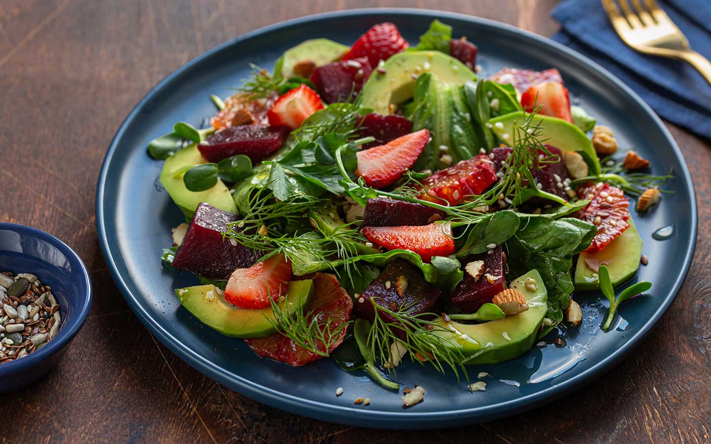 A colorful salad with avocado, beet, strawberries, and greens on a blue plate, with a bowl of seeds nearby.
