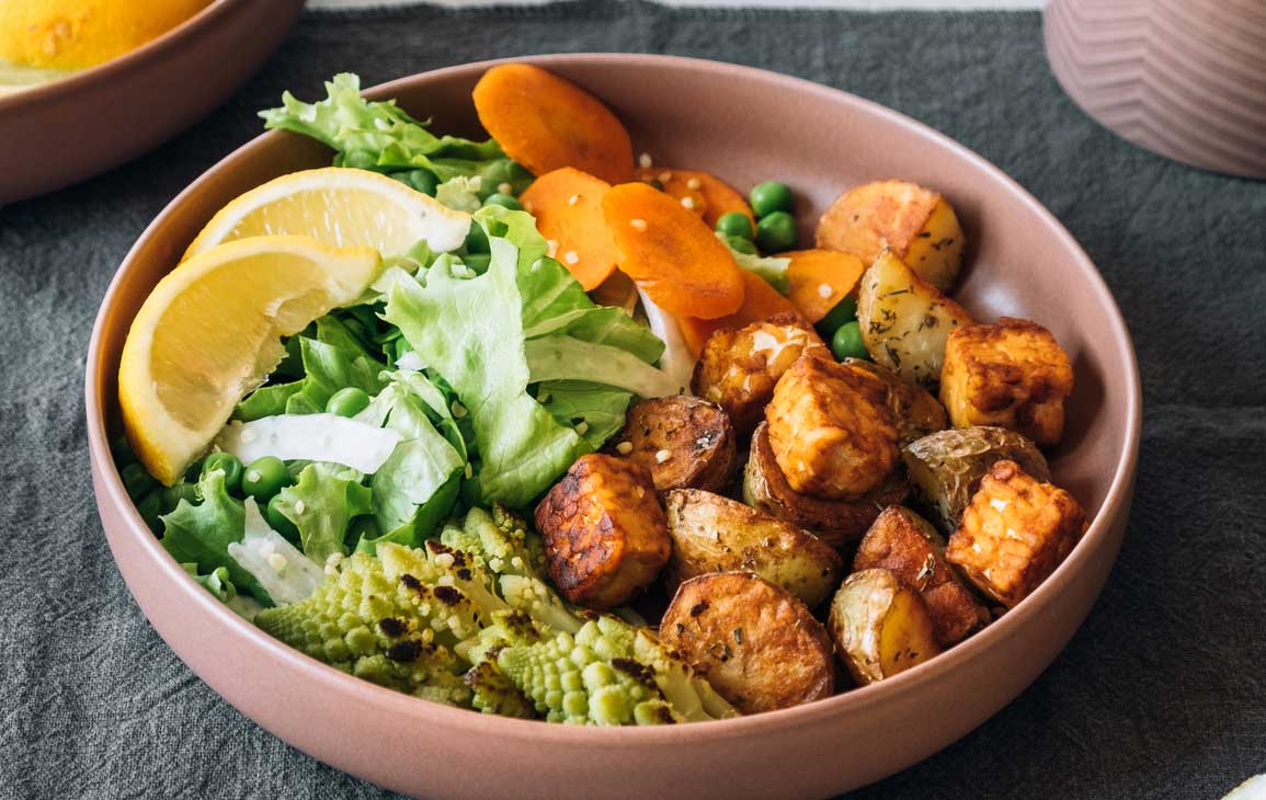 A bowl of mixed salad with lettuce, lemon slices, green peas, tofu, roasted potatoes, carrots, and Romanesco broccoli.