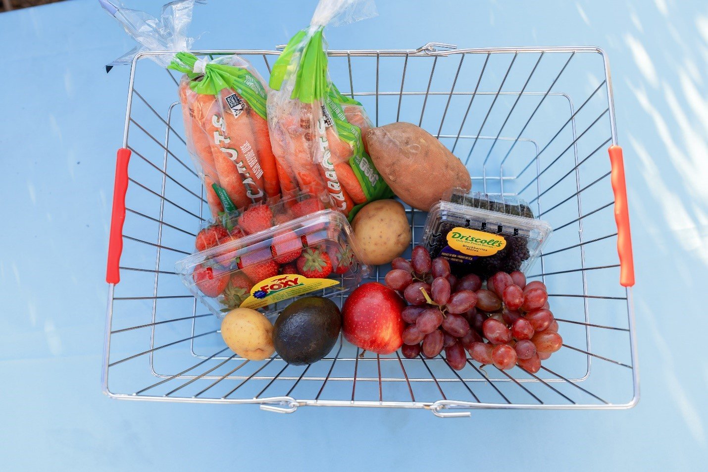 A metal basket containing carrots, sweet potato, strawberries, blackberries, grapes, apple, potato, and avocado.