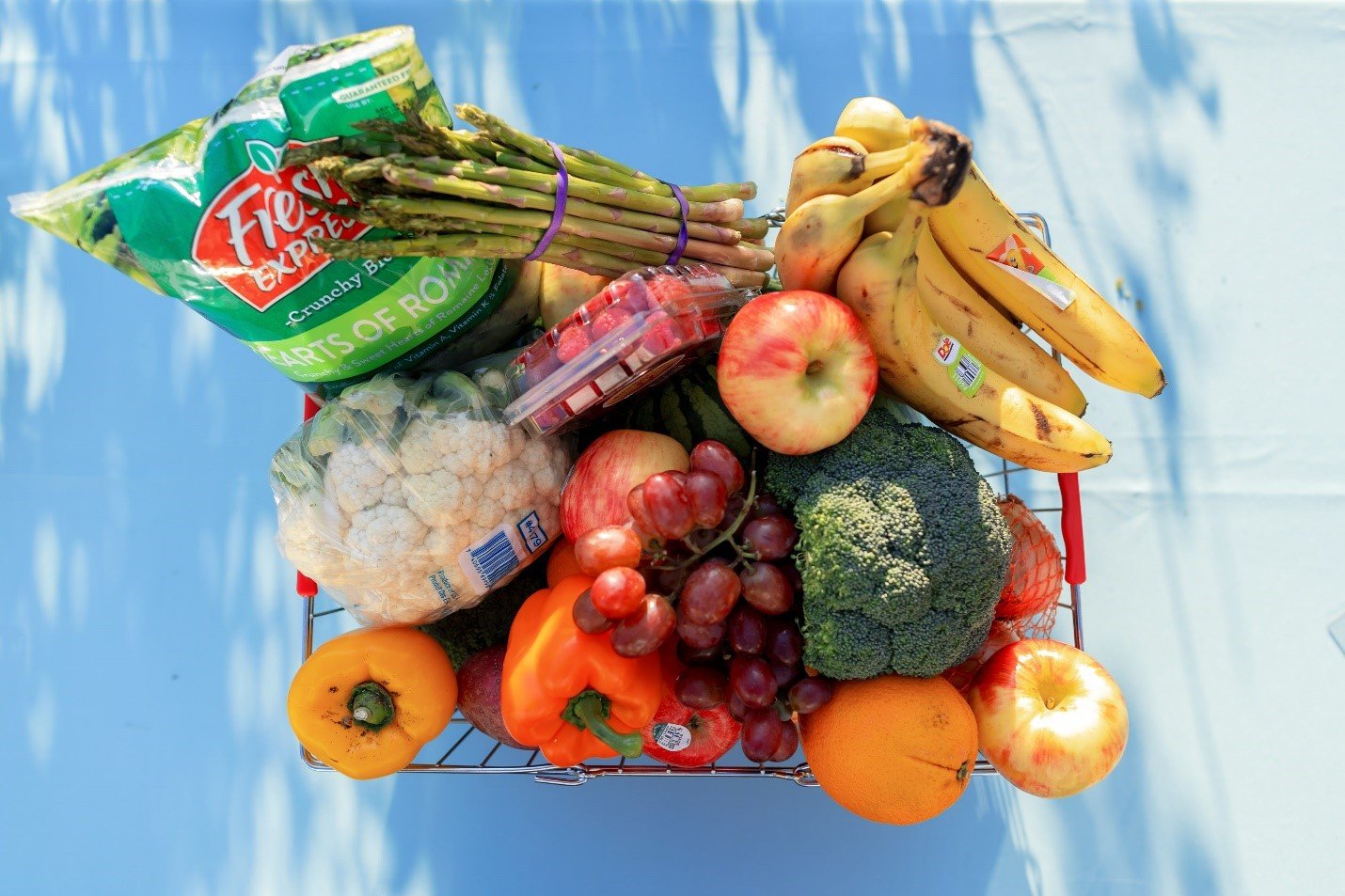 Overhead view of a grocery cart filled with assorted fruits and vegetables including bananas, apples, broccoli, cauliflower, grapes, oranges, bell peppers, and a bag of romaine lettuce.