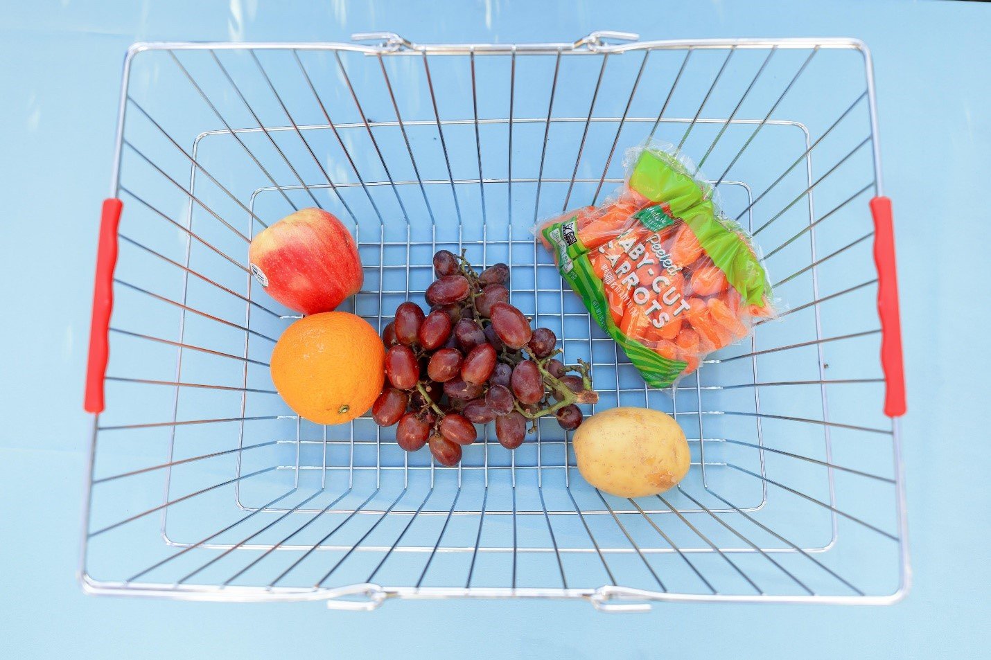 Metal shopping basket with fruits, vegetables, and a bag of carrots against a light blue background.