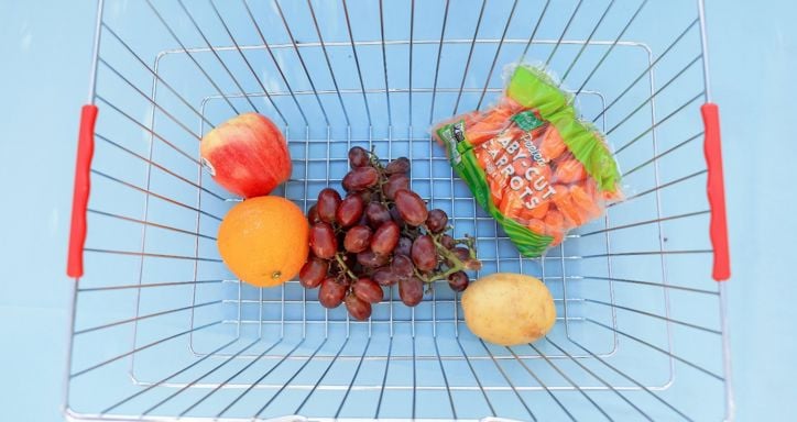 Metal shopping basket with fruits, vegetables, and a bag of carrots against a light blue background.