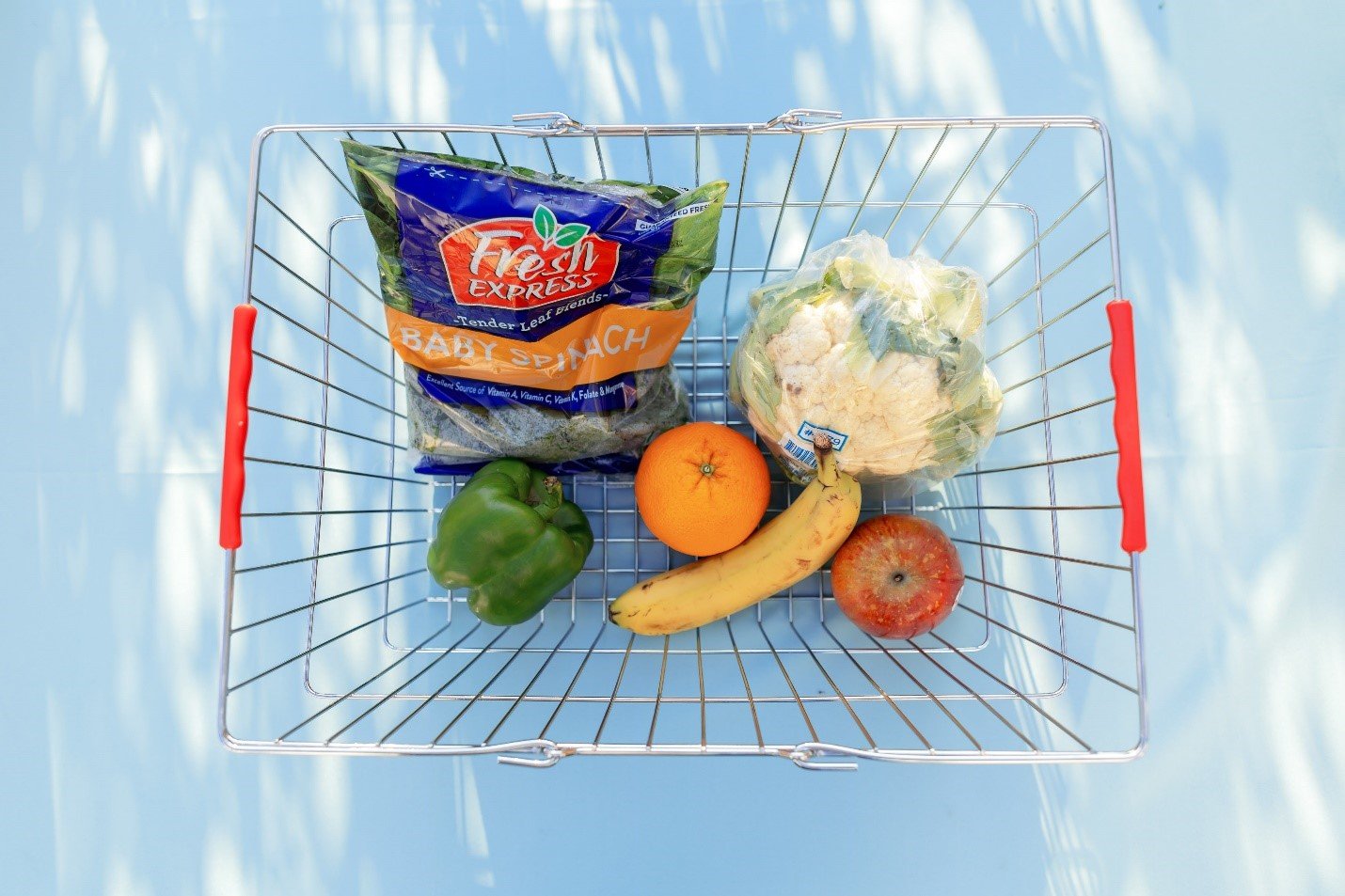 A grocery basket containing Fresh Express Baby Spinach, a green pepper, an orange, bananas, an apple, and a cauliflower.