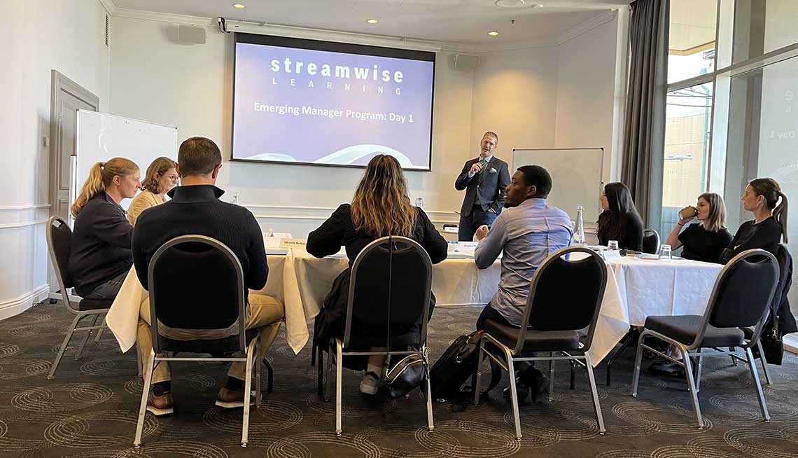 A group of people sits at tables arranged in a U-shape, facing a presenter who stands near a screen displaying "Streamwise Learning - Emerging Manager Program: Day 1". The room is well-lit with large windows and a whiteboard on the side.