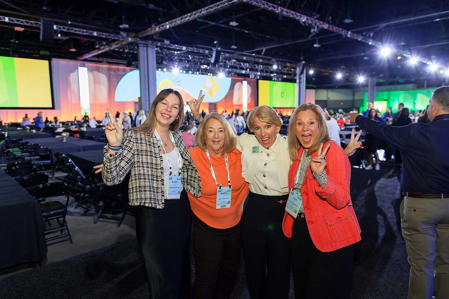 Four women including Cathy burns smiling and posing with peace signs at an indoor event with bright lighting and a large screen in the background. Four women including Cathy burns smiling and posing with peace signs at an indoor event with bright lighting and a large screen in the background.