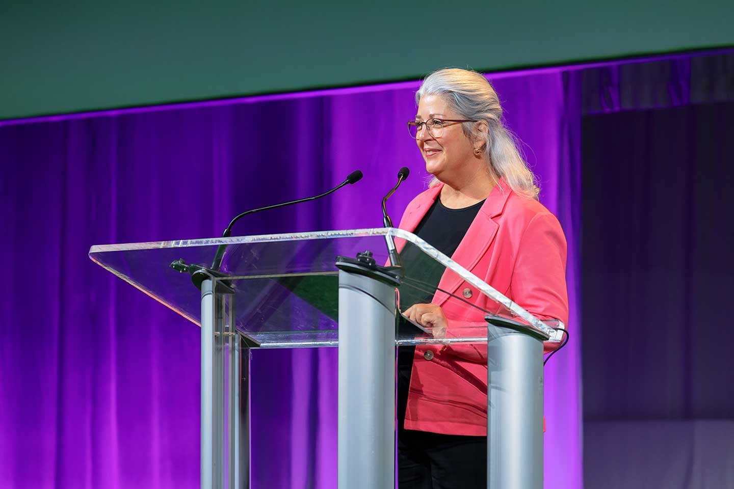 Brenda Briggs in a pink blazer speaks at a clear podium with a purple-lit background. Brenda Briggs in a pink blazer speaks at a clear podium with a purple-lit background.