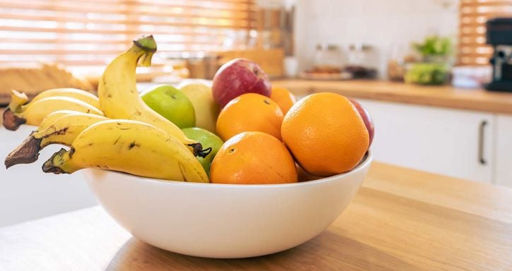 A bowl of bananas, apples, and oranges on a kitchen countertop with sunlight through wooden blinds.