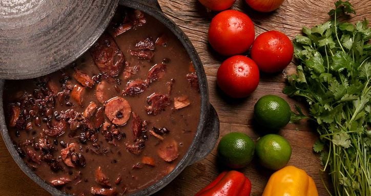 A clay pot with a stew of black beans and sausage, surrounded by fresh vegetables on a wooden surface.