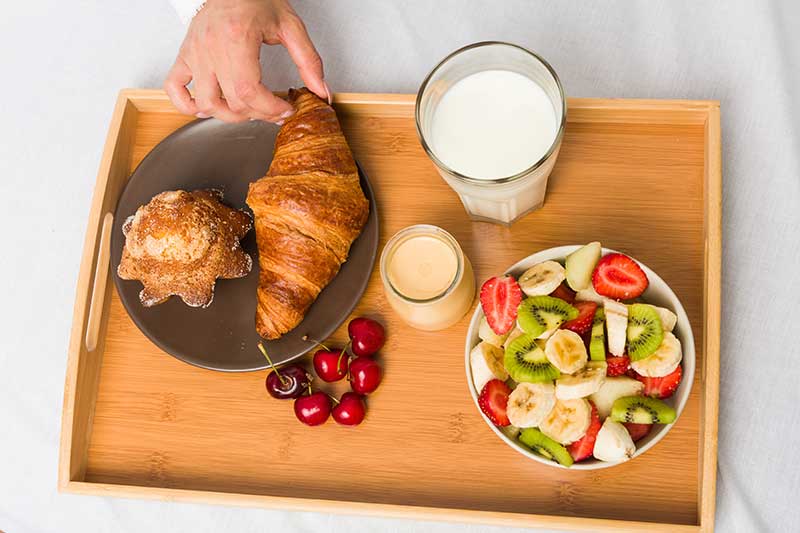 A breakfast tray with a croissant, muffin, cherries, a glass of milk, a jar of yogurt, and a bowl of mixed fruit.