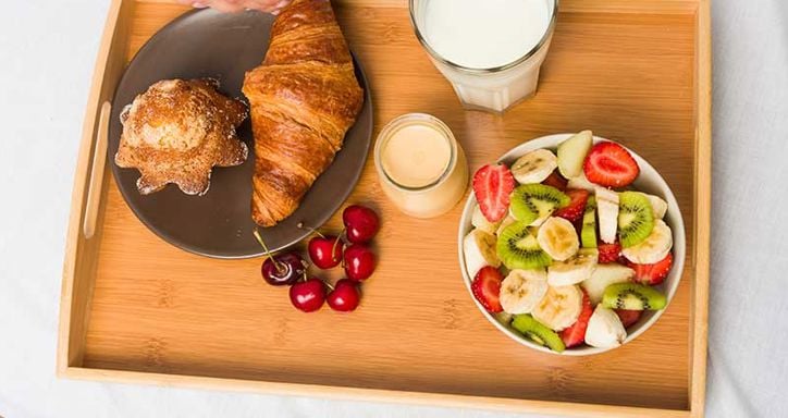 A breakfast tray with a croissant, muffin, cherries, a glass of milk, a jar of yogurt, and a bowl of mixed fruit.