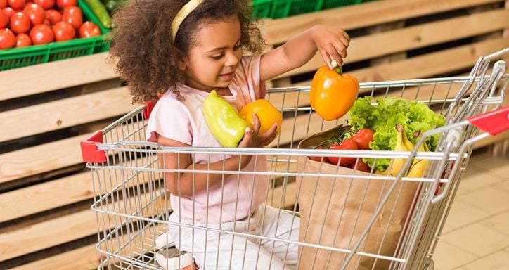 Young child in a shopping cart holding bell peppers in a grocery store.