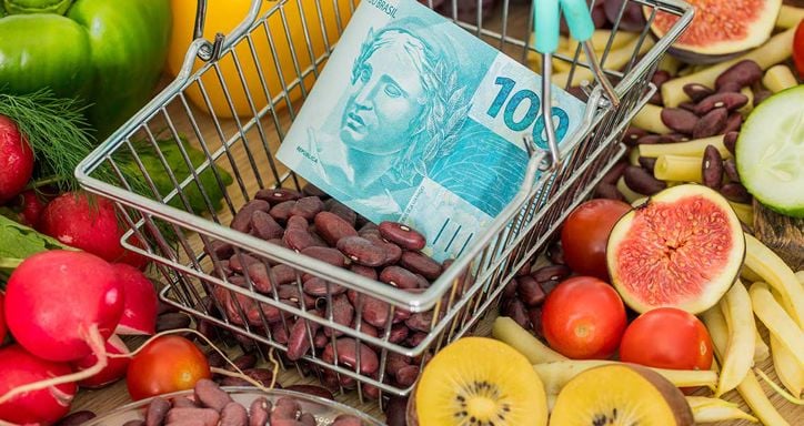 A shopping basket with red kidney beans and a 100 Brazilian real banknote, surrounded by fresh fruits and vegetables.