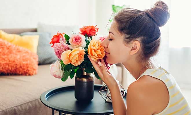 A woman in her living room gently holds a peach colored flower in her hand and smells the flower with her eyes closed.