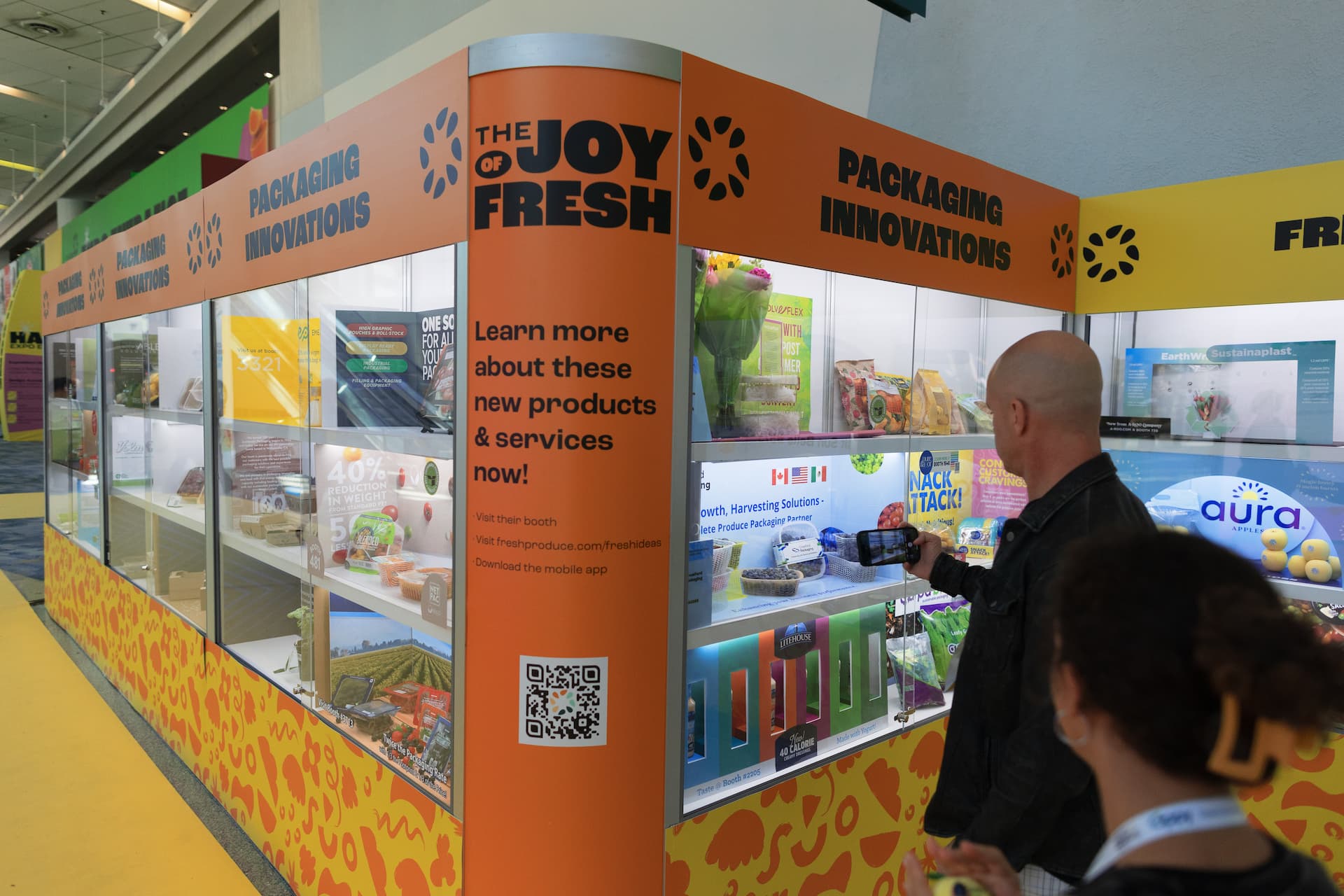 A man takes a picture of a bright orange and yellow display at Fresh Ideas Showcase 2023. The display has the words The Joy of Fresh in very large black letters.