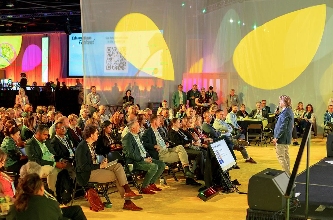 A speaker addresses an audience during the educational festival at the Global Produce and Floral Show, with colorful abstract projections in the background.