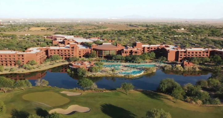 Aerial view of a desert resort complex with reddish-brown buildings, turquoise pools, and surrounding greenery.