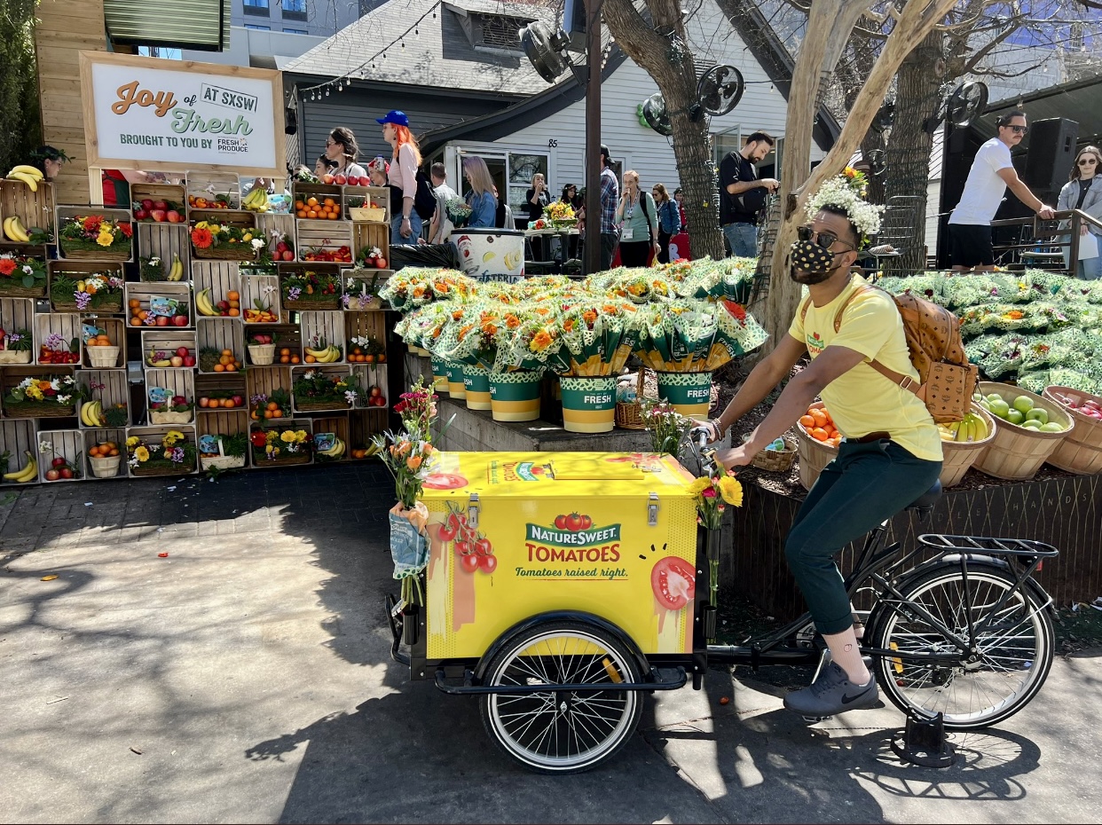 A gentleman with flowers in his hair rides a vendor style bike giving out nature sweet tomatos.