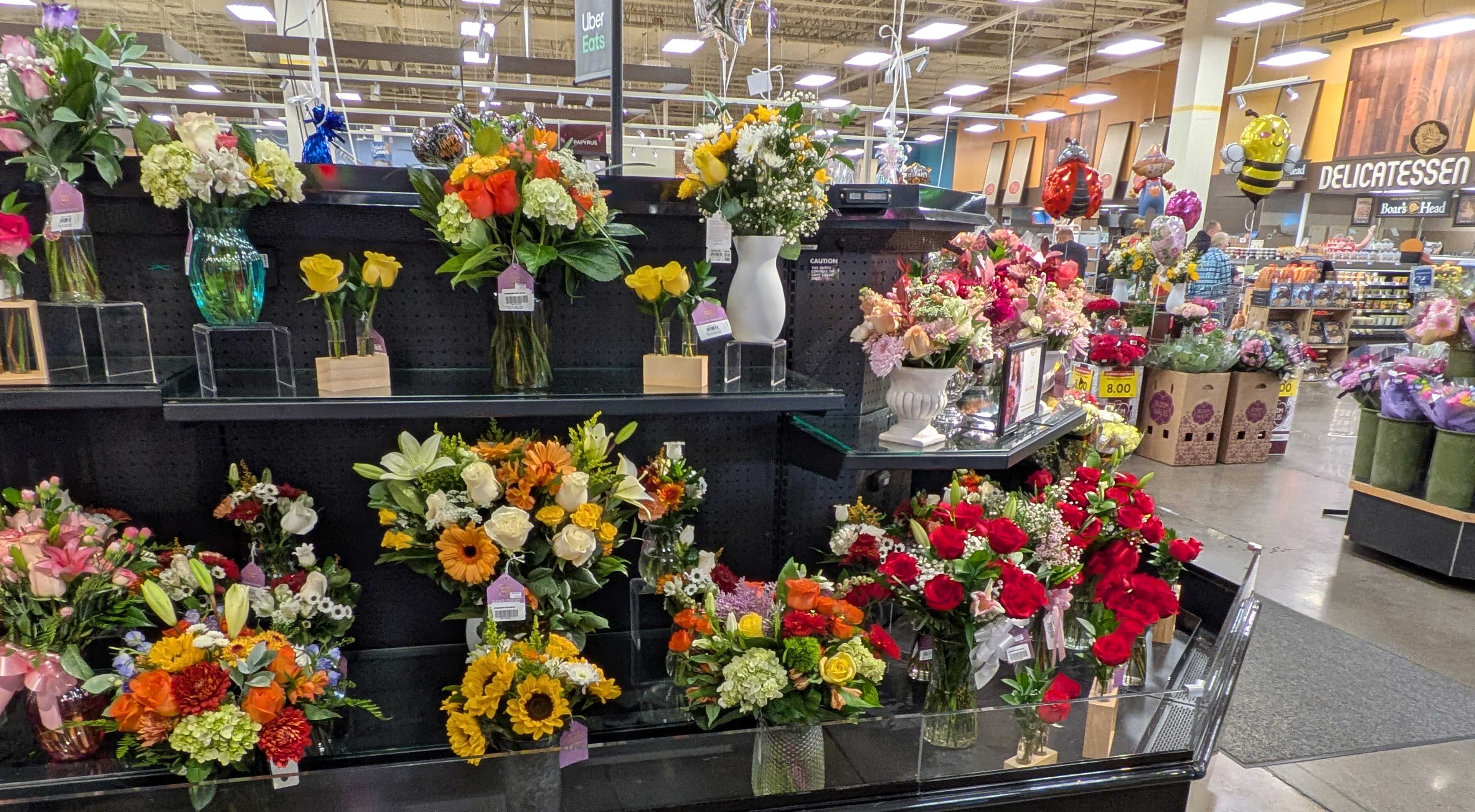 A grocery store flower display with various colorful bouquets of roses, lilies, and daisies in clear vases, along with decorative balloons in the background.