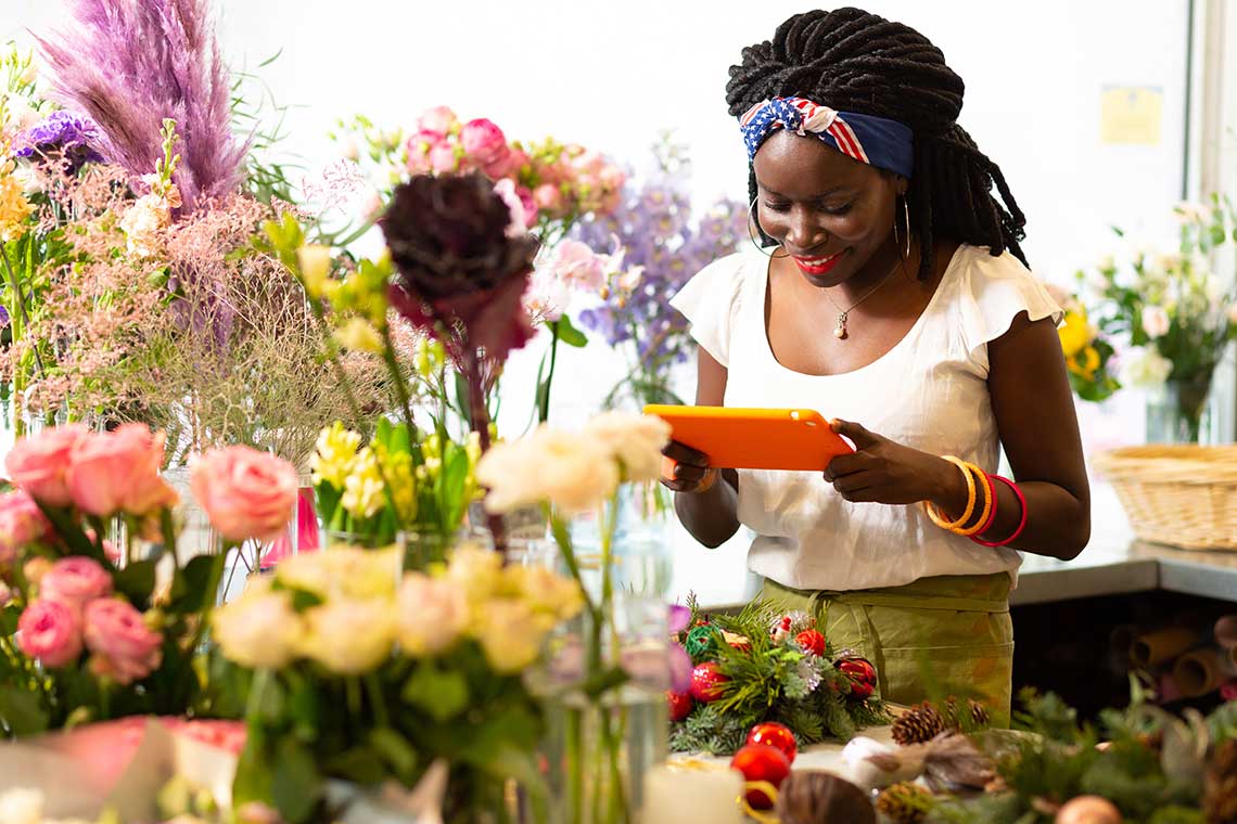A woman holding an orange tablet among colorful flowers in a shop.