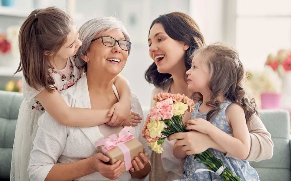 A grandmother, mother, and daughter share a joyful moment together, with the mother holding a bouquet of flowers. The image captures a warm, familial celebration.