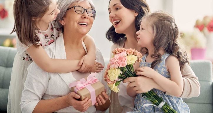 A grandmother, mother, and daughter share a joyful moment together, with the mother holding a bouquet of flowers. The image captures a warm, familial celebration.