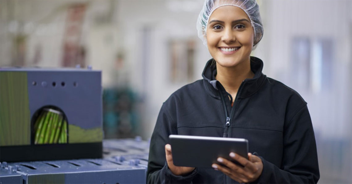 Smiling woman wearing a hair net and holding a tablet stands next to boxes of asparagus.