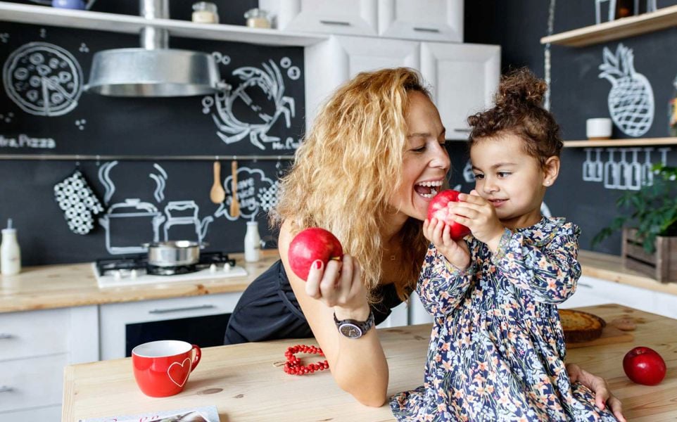 Mother and daughter hold bright red apples while daughter sits on the counter in the kitchen.