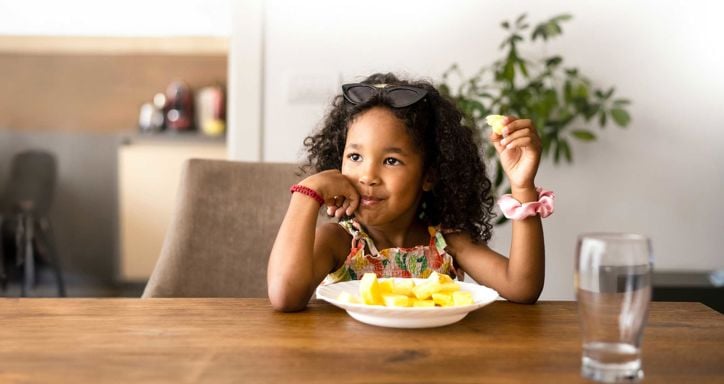 A small child with sunglasses resting on her head sits at a long wooden dining table eating a bowl of fruit.