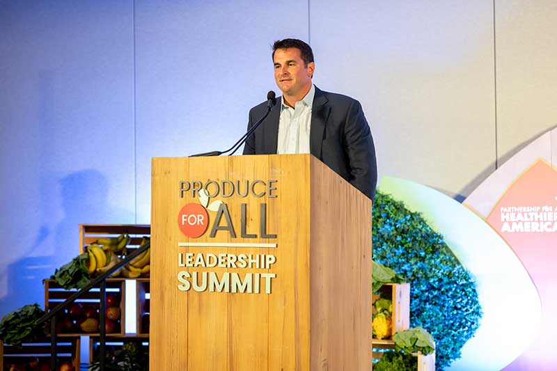   Alex DiNovo  Chair  President & CEO  DNO Produce standing behind a podium at the Produce for All Leadership Summit. The background features a display of fresh produce and the logo of Partnership for a Healthier America.