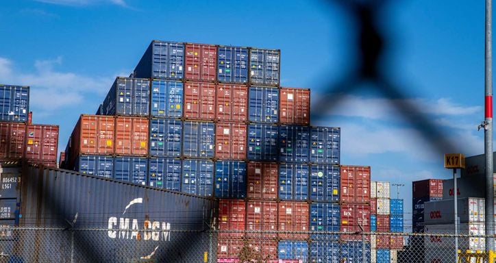 A stack of red, blue, and white shipping containers in a yard, seen behind a blurry chain-link fence, under a blue sky.