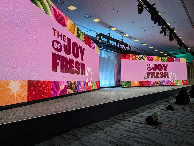Stage with two large screens displaying "THE JOY OF FRESH" in bold letters against a pink background, surrounded by colorful fruit and flower imagery.