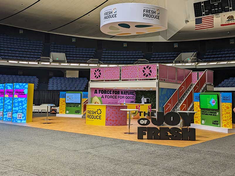 Colorful exhibition booth in an arena with multiple displays and a sign reading "THE JOY OF FRESH."