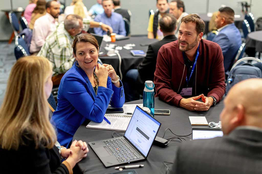 Attendees converse around a conference table with an open laptop, notebooks, phones, and a water bottle.