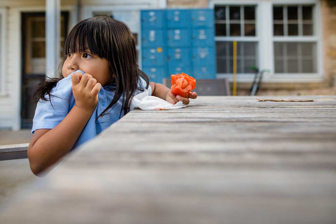 Child at a wooden table holding red fruit with blue lockers in the background.