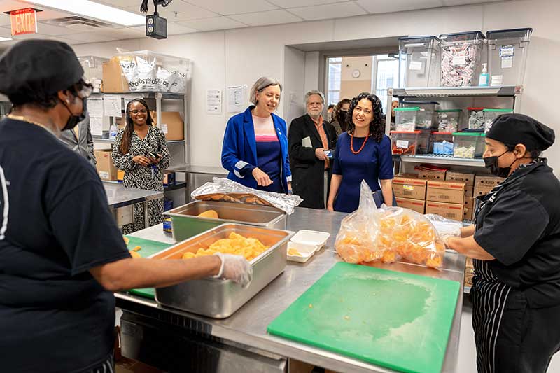 USDA Deputy Secretary Xochitl Torres Small and Deputy Under Secretary Stacy Dean greet women from DC Central Kitchen as they prepare vegetables in a school cafeteria kitchen.