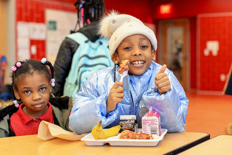 Two children sit at a lunch table in a school cafeteria where one child gives a thumbs up signal as she eats strawberry French toast. She has a banana, orange juice, and milk on her tray as well.