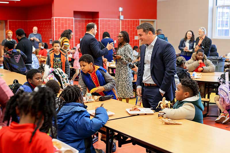 Foundation for Fresh Produce Board Chair and President of DNO Produce, Alex DiNovo, visits with children at a school cafeteria table. The children are eating breakfast and people are socializing in the school cafeteria.