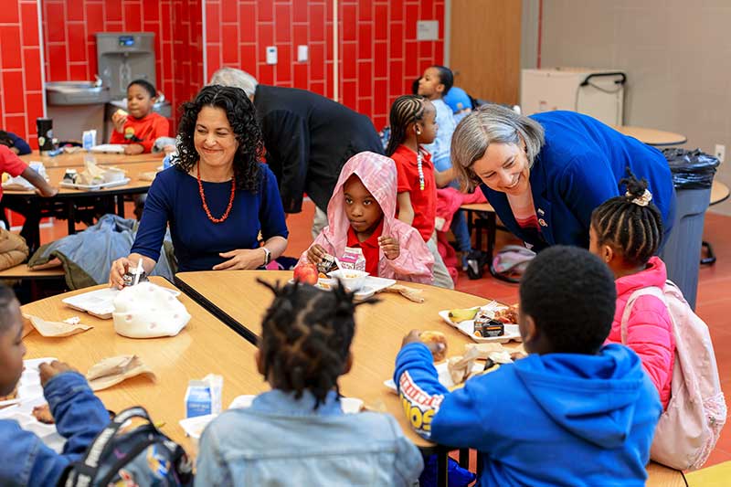 USDA Deputy Secretary Xochitl Torres Small and Deputy Under Secretary Stacy Dean smile as they visit with children at a lunch table in a school cafeteria. The children are eating breakfast, including the strawberry French toast prepared by DC Central Kitchen.