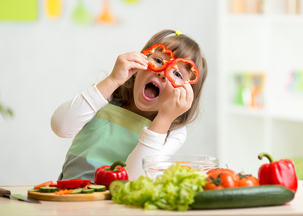 Girl having fun with food vegetables in the kitchen by holding up two slices of red pepper up as eyeglasses.