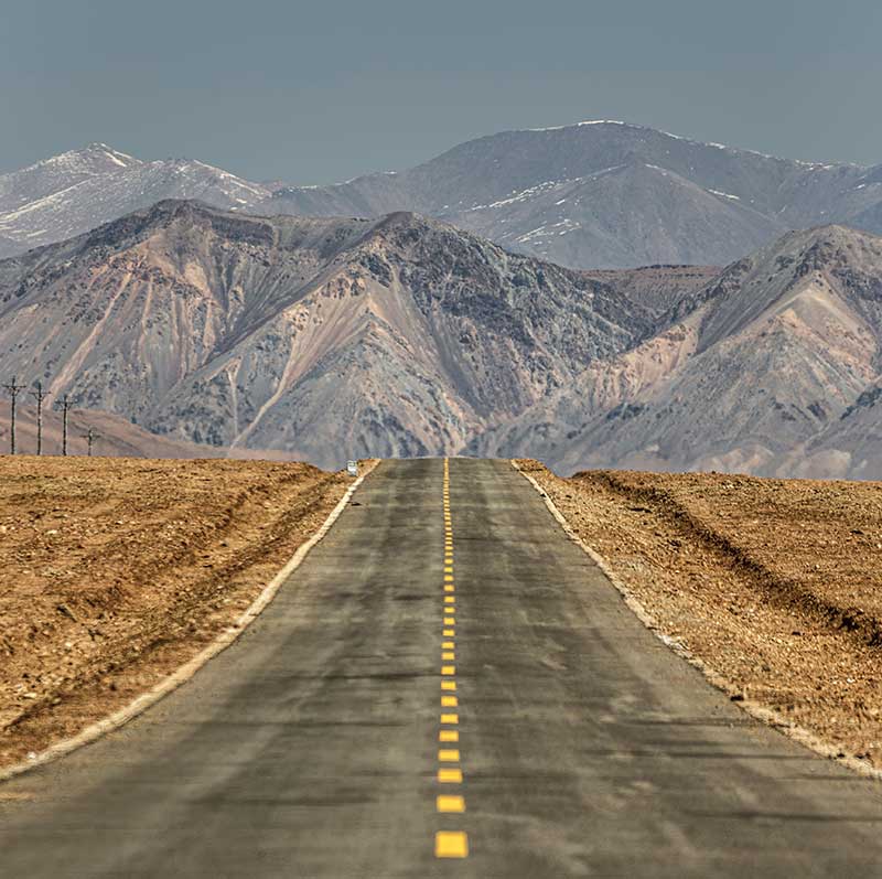 A straight road leading through a barren desert landscape towards distant mountains under a clear blue sky.