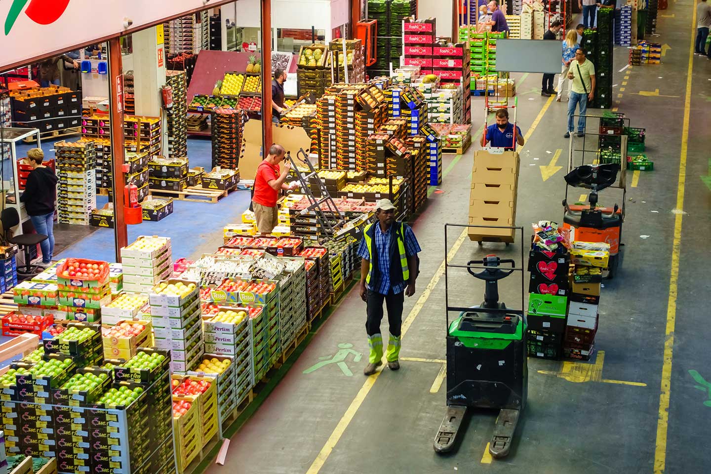 A busy wholesale market filled with stacked fruit boxes. Workers are engaged in tasks, with forklifts visible on the floor and brightly colored produce on display.
