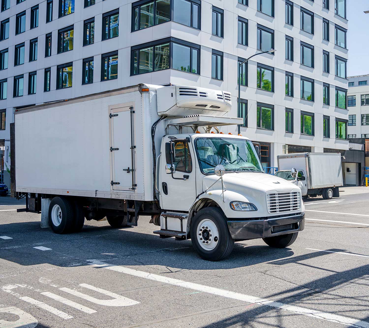 White refrigerated box truck parked on a city street beside a modern windowed building.