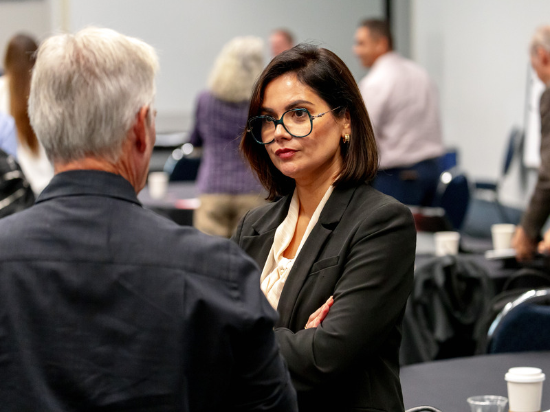 A woman with glasses in a blazer talking to a man at a professional event.