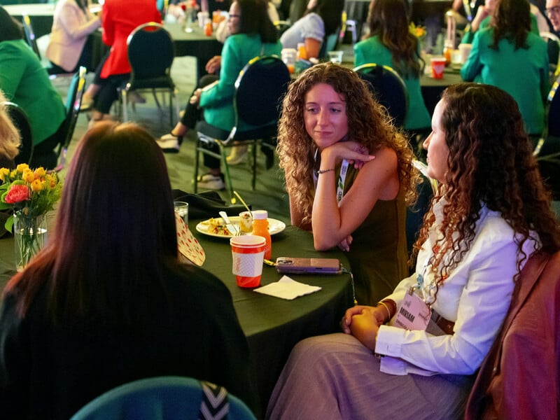 People seated around tables at a social event, with three women in conversation in the foreground.