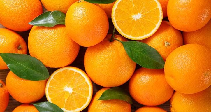 A pile of fresh oranges with two cut in half, showing the interior, and some green leaves scattered among them.