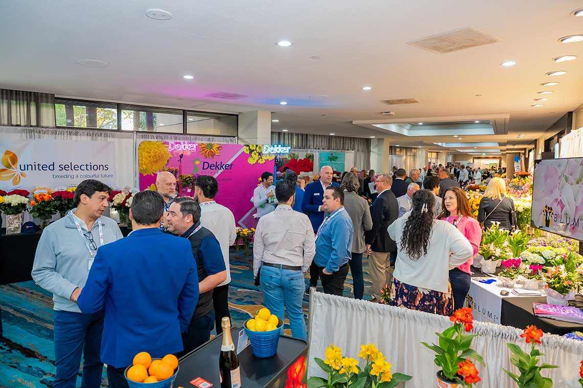 A bustling trade show with people talking near colorful flower displays and brand banners.